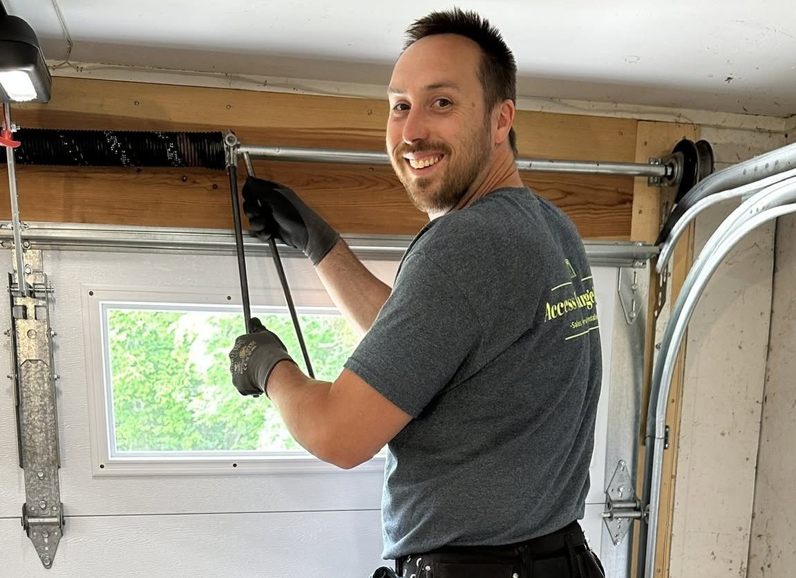 Man wearing gloves and a gray t-shirt tightens a garage door spring while standing in a residential garage, smiling at the camera.