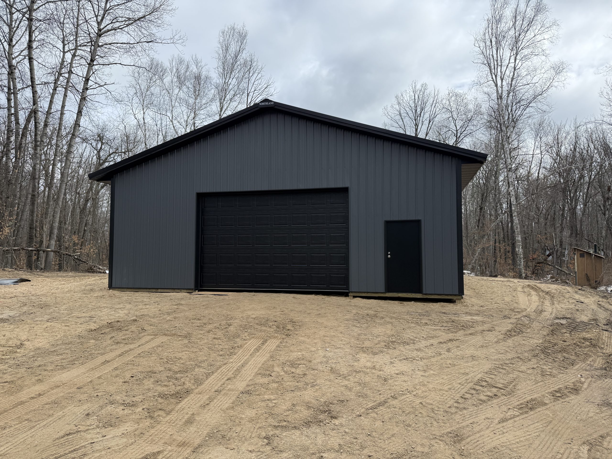 Gray metal garage with a large dark door, set in a leafless forest under a cloudy sky.