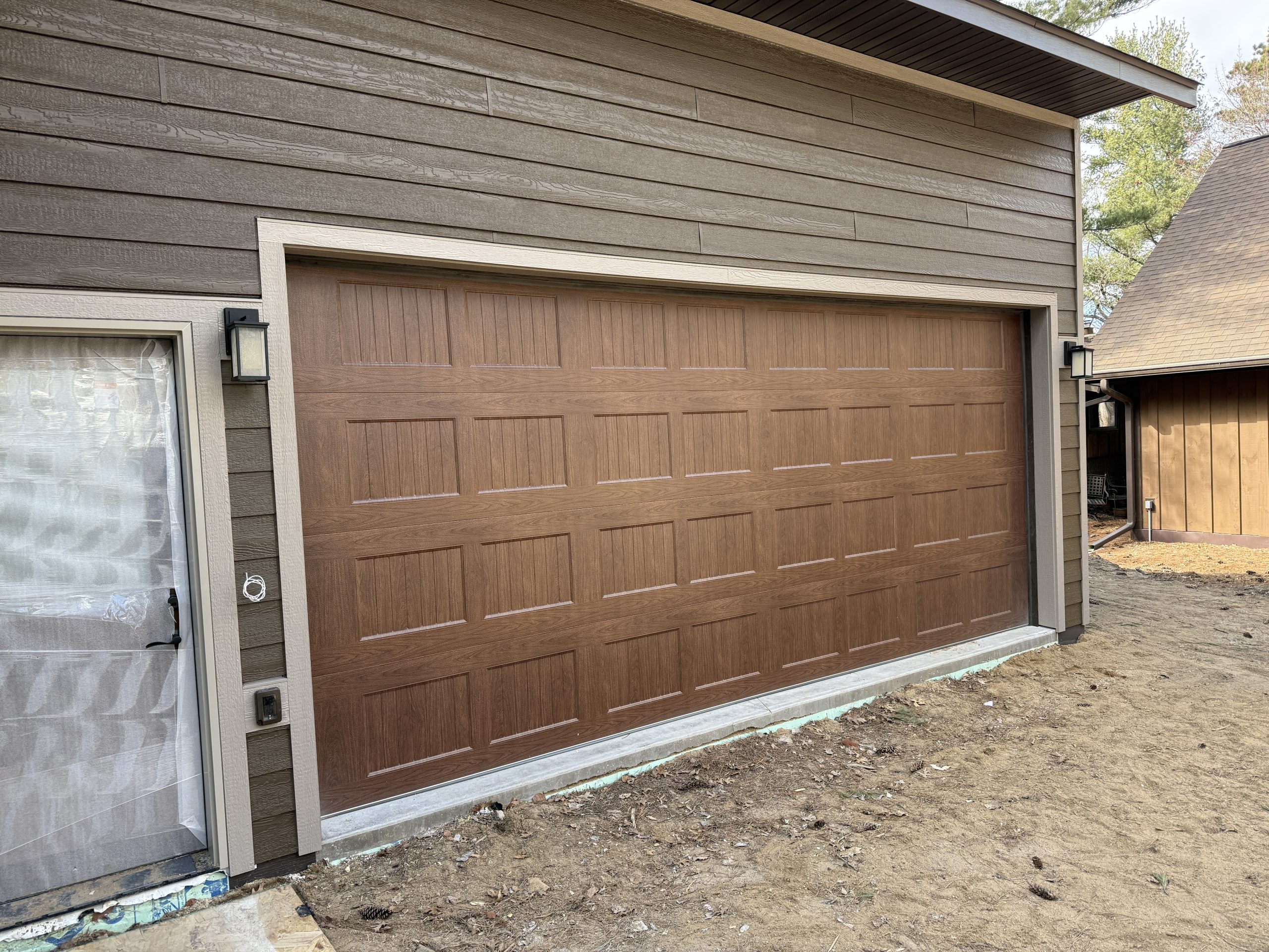 Brown sectional garage door with beige trim on a newly sided house, outdoor lights mounted on either side, and a dirt yard in front (under construction).