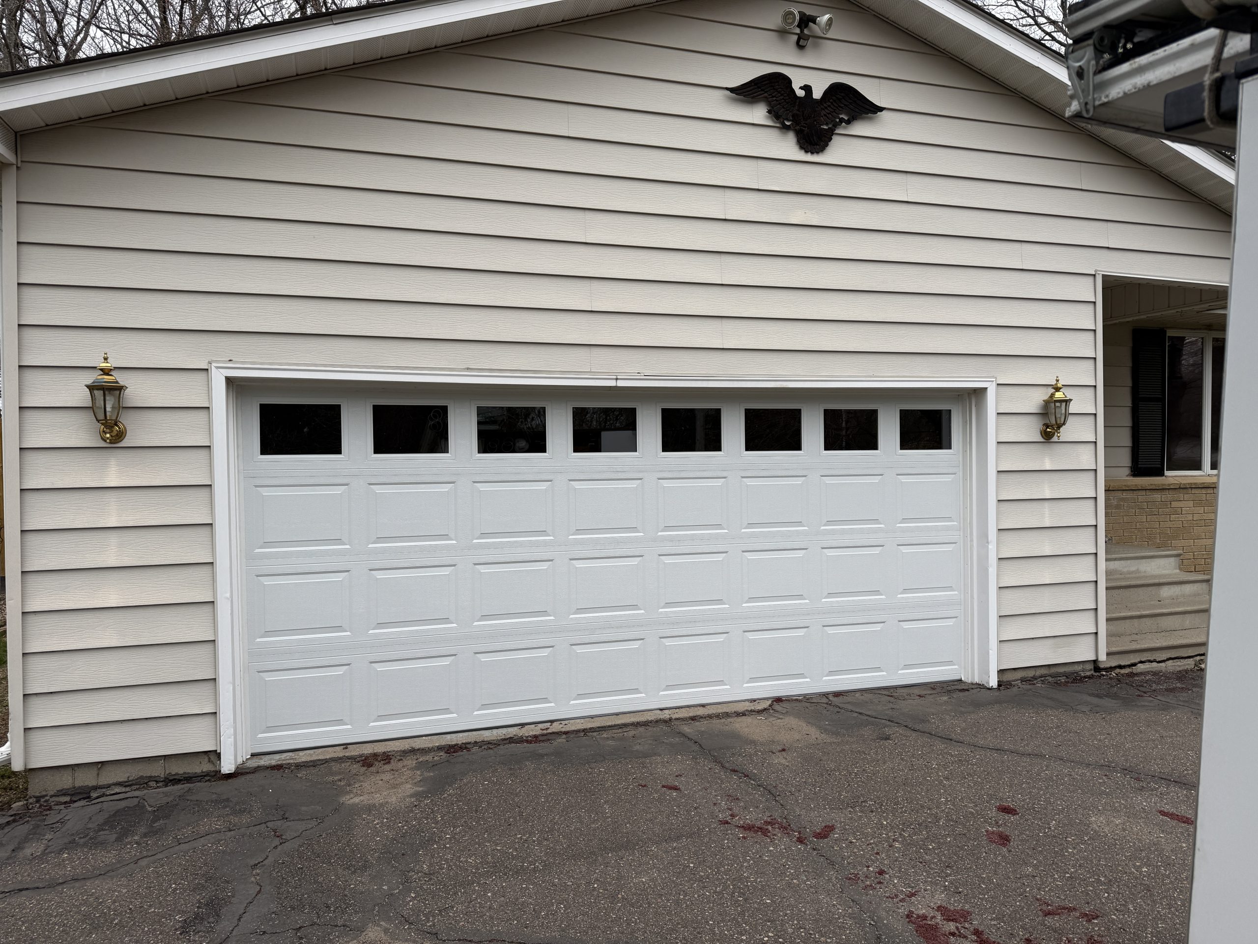 Front of a beige-siding house with a white paneled garage door, a black eagle crest above, and gold wall lanterns on both sides.