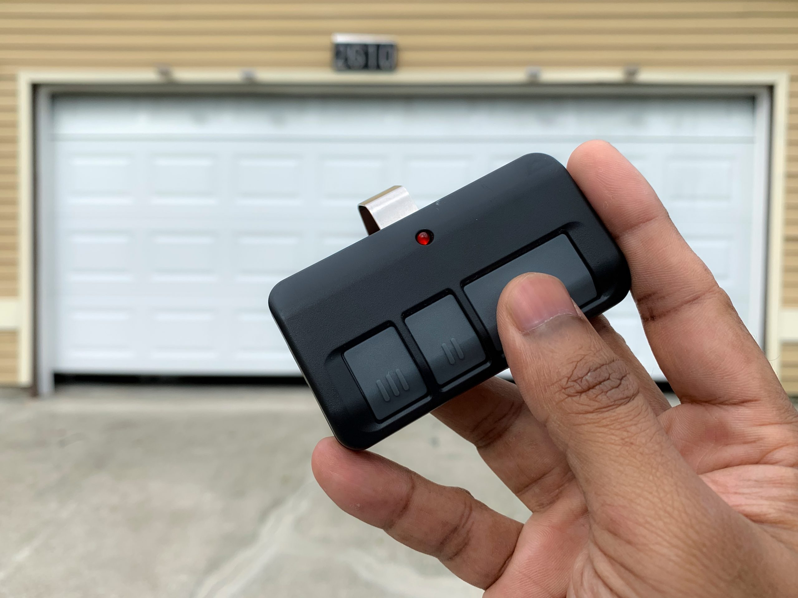 Close-up of a hand holding a black garage door remote with a red indicator light, a blurred white garage door in the background.