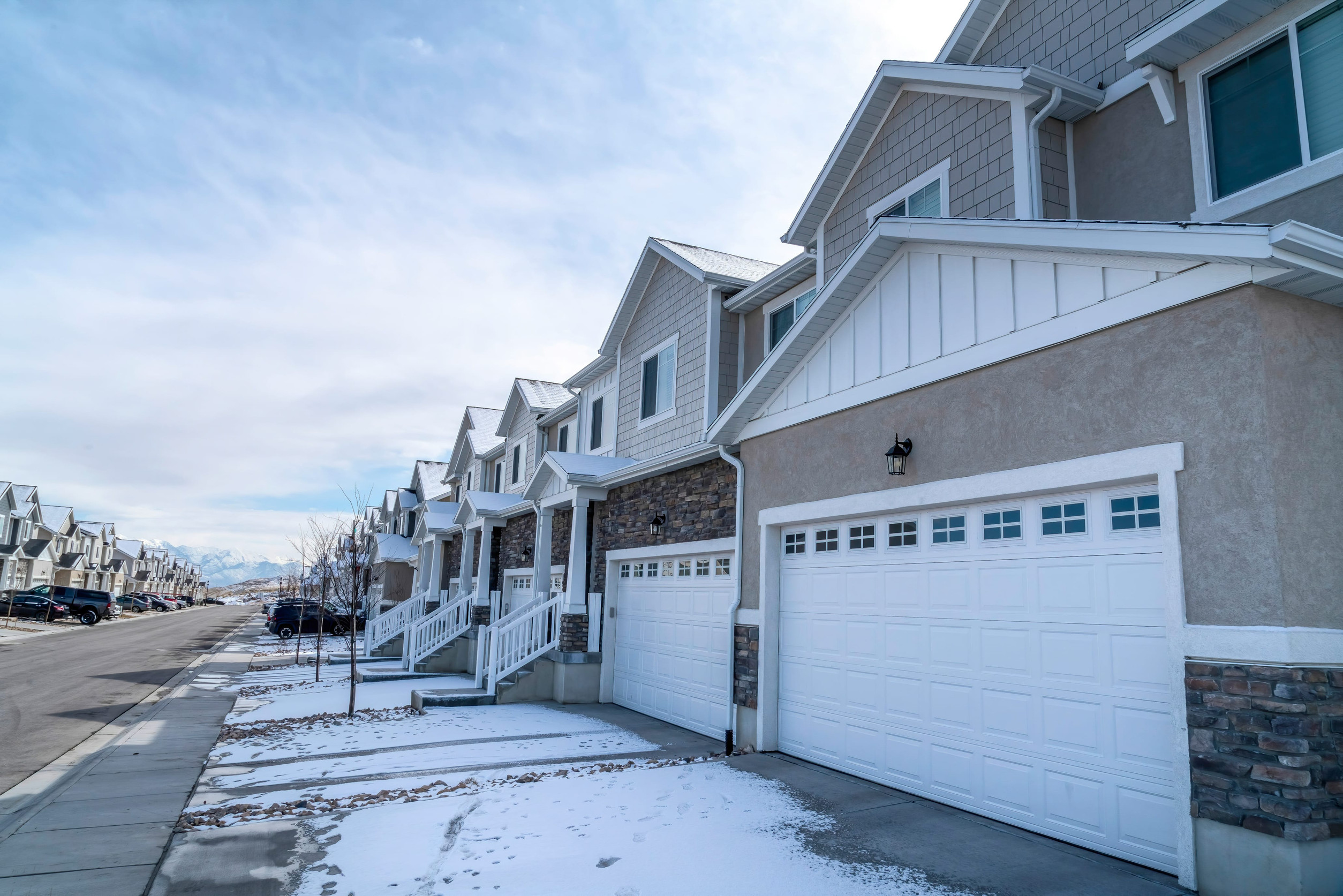 Row of beige townhouses with white garage doors along a snowy street under a blue sky, distant mountains visible to the left.
