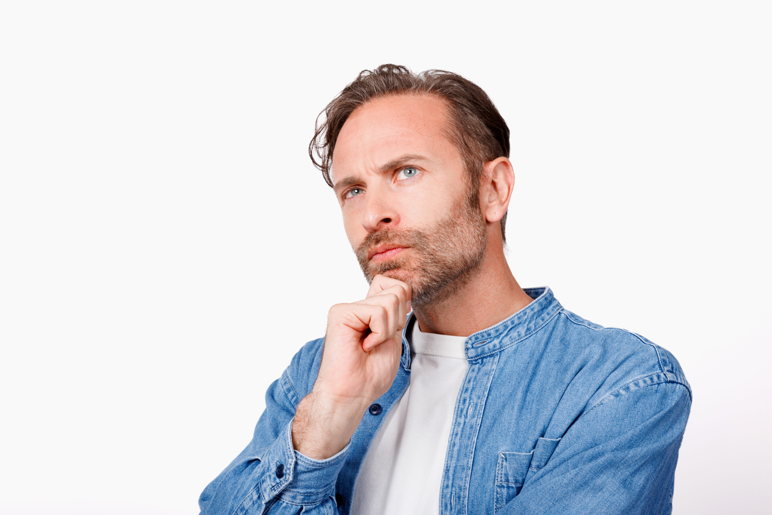 Thoughtful man with light beard in a blue denim shirt, resting his chin on his hand and looking upward.