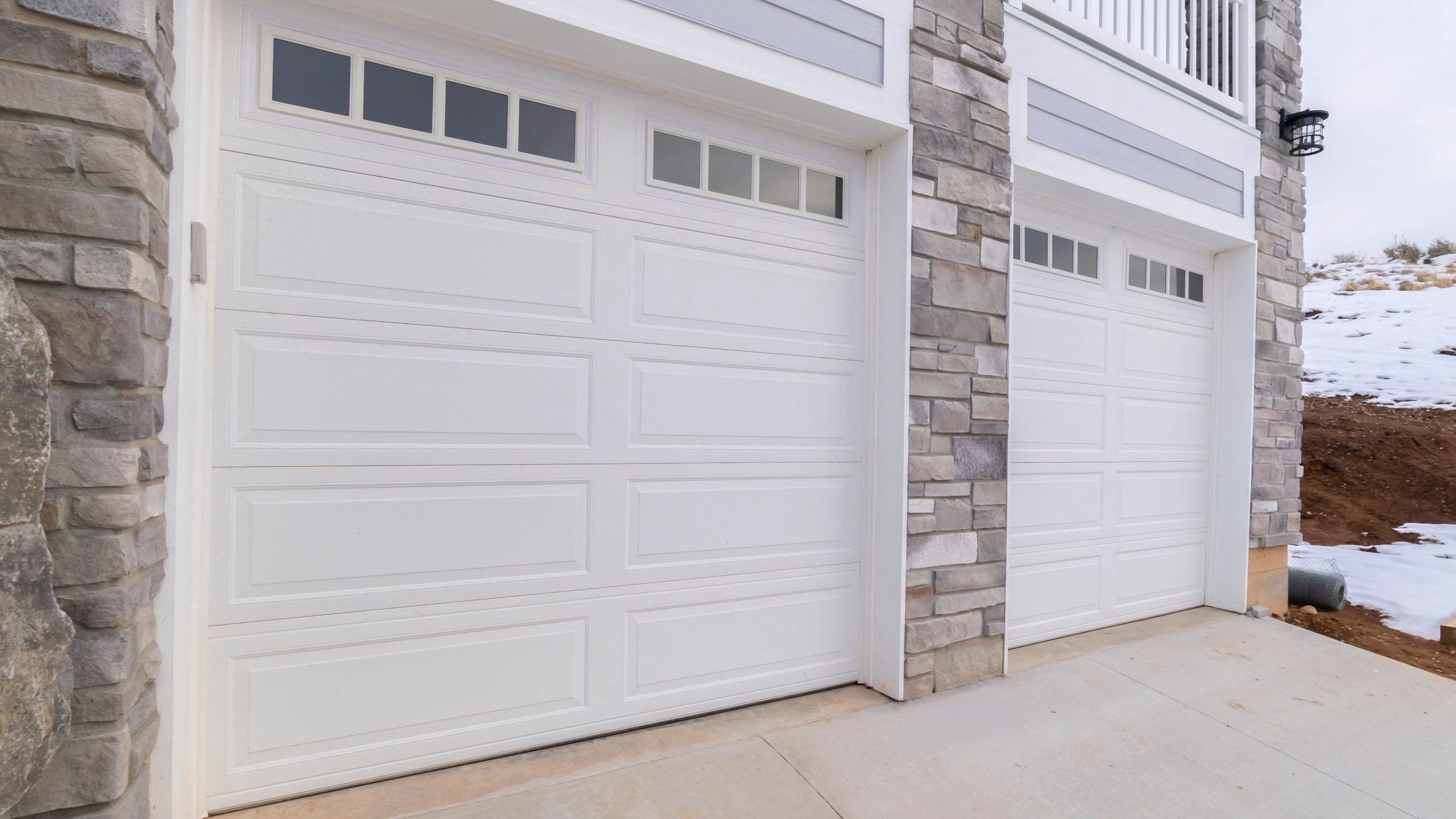 Three white garage doors with small rectangular windows at the top, flanked by stone columns and a snowy landscape nearby.