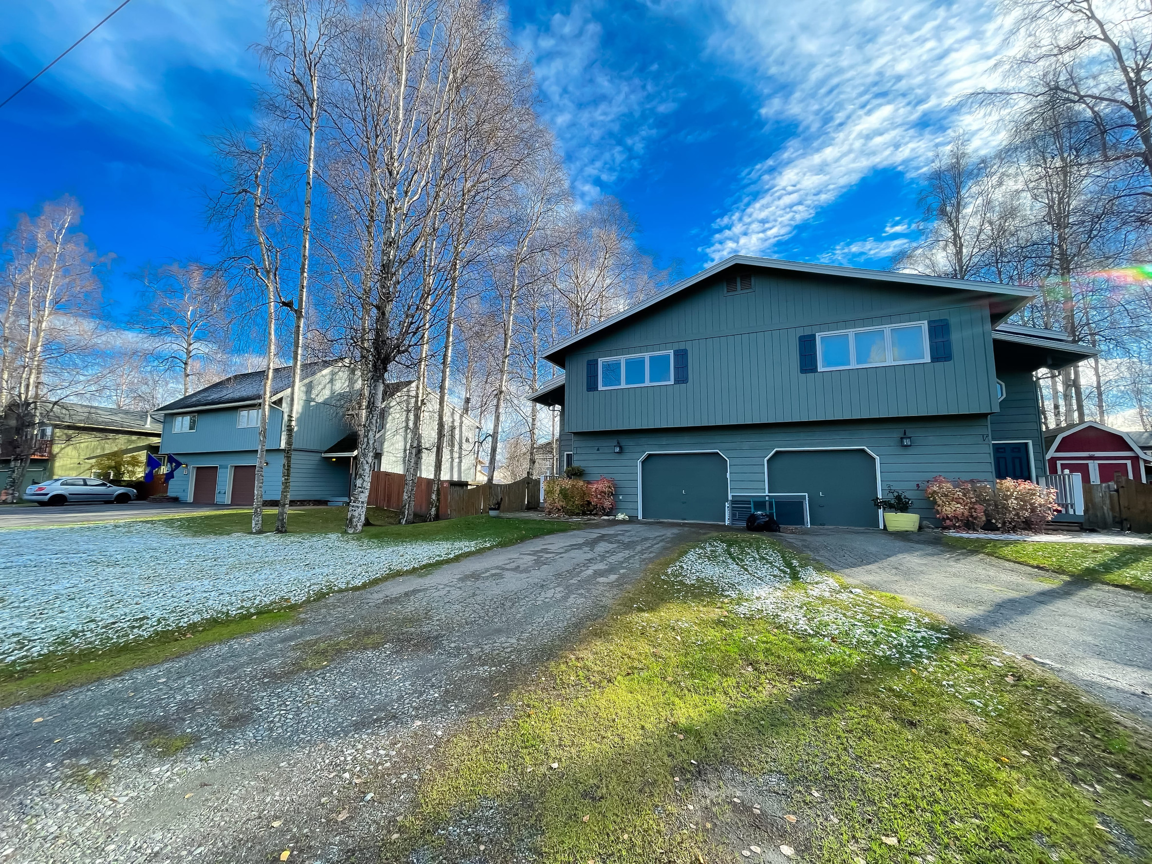 Suburban street with leafless trees, a teal two‑story house, and a bright blue sky with clouds.